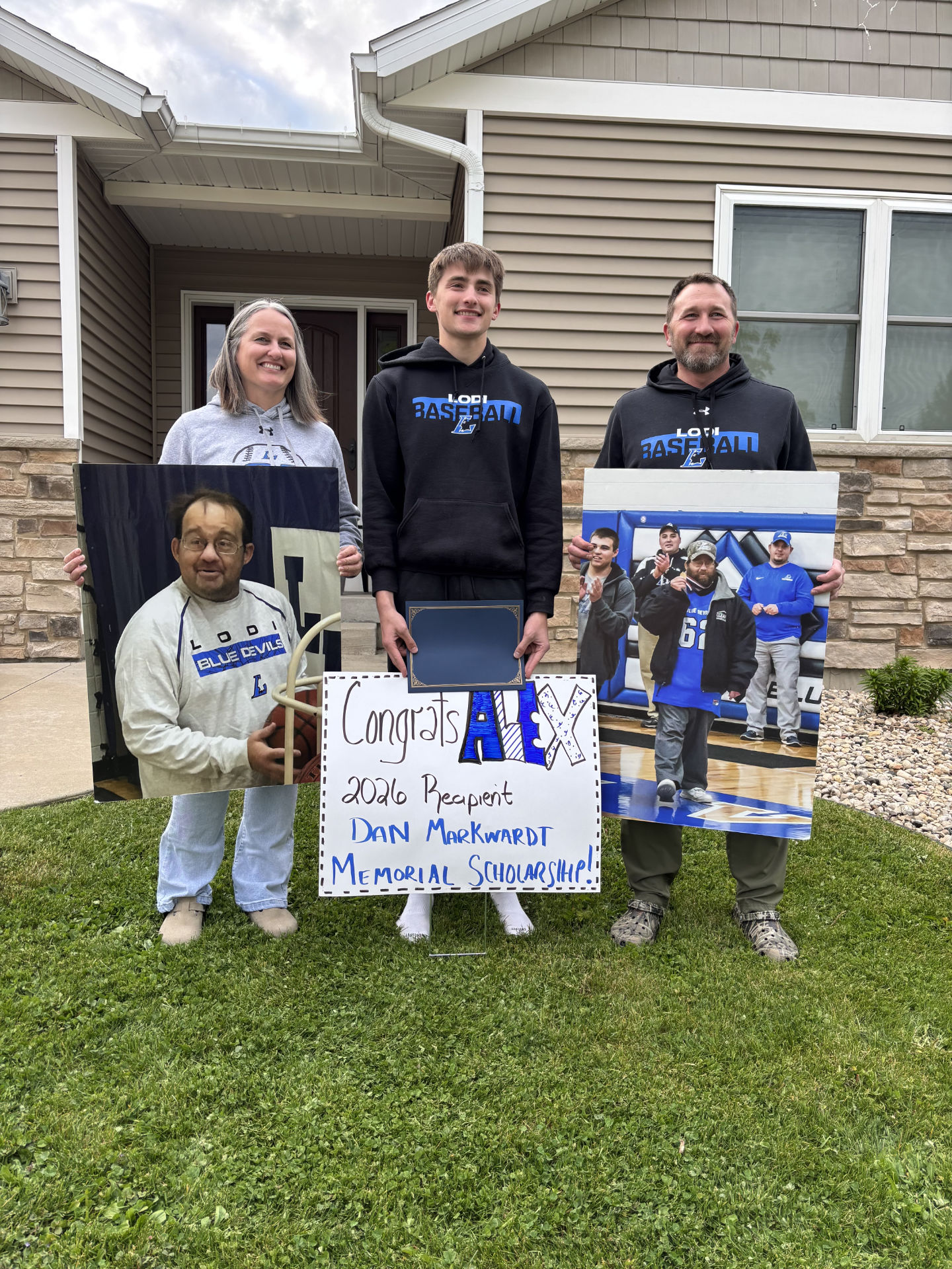 Alex Breunig with his parents, Rachel and Landon, holds a scholarship. Signs are displaying a congratulations sign and pictures of Dan Markquart.