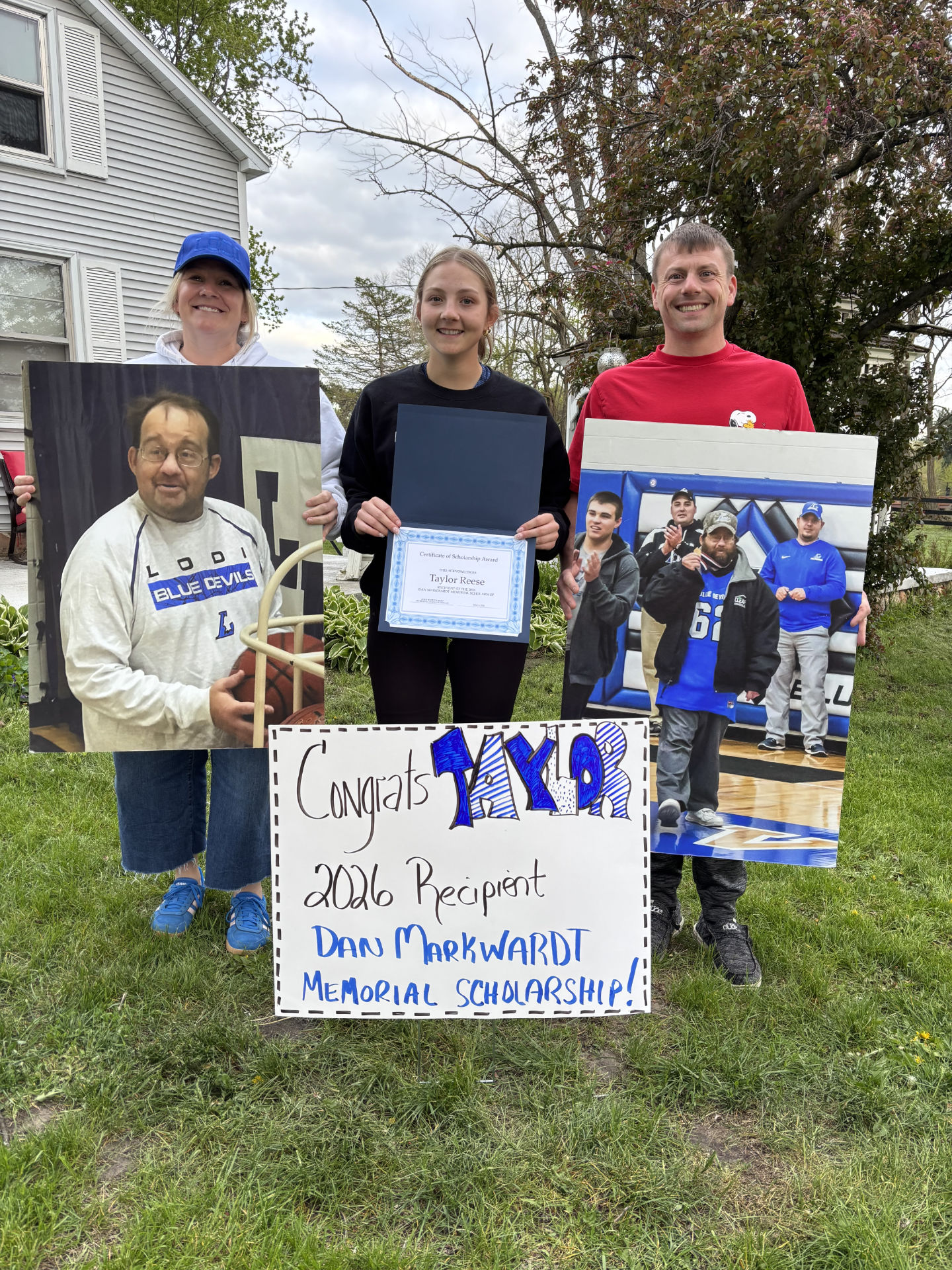 Taylor Reese with her parents, Andy and Samantha, holds a scholarship. Signs are displaying a congratulations sign and pictures of Dan Markquart.